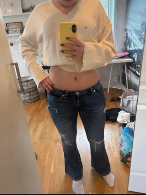 Mörkblå bootcut jeans med slitningar - Säljer ett par mörkblå bootcut jeans med snygga slitningar och detaljerade bakfickor. Jeansen har låg midja och klassisk femficksdesign. Perfekta för dig som gillar en avslappnad och trendig look.