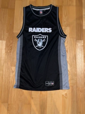 Raiders linne från Primark NFL - Snyggt svart och grått Raiders linne från Primark, officiellt NFL-merch. Stort Raiders-tryck och logga framtill, samt NFL-märke i nacken. Perfekt för dig som älskar amerikansk fotboll och vill ha en sportig look.