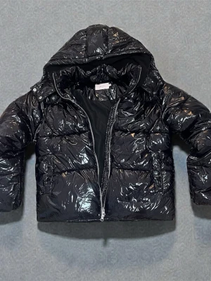 Svart glansig pufferjacka från Nelly - Säljer en svart, glansig pufferjacka från Nelly i storlek M. sopass oversized look, med huva och dragkedja framtill. Yttermaterialet är 100% polyamid och fodret är återvunnen polyester. Perfekt för kalla dagar när du vill ha en cool och trendig vibe.