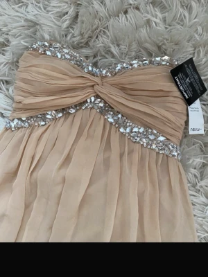 Champagnefärgad bandeauklänning Nelly - Champagnefärgad bandeauklänning från Nelly med glittriga paljettdetaljer längs bysten och omlottliknande twist framtill. Klänningen har ett luftigt, lager-på-lager fall och är tillverkad i ett lätt, skirt material. Perfekt för bal eller festliga tillfällen.