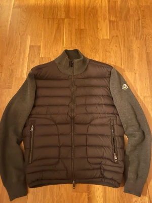 Grå Moncler pufferjacka med stickade ärmar - Säljer en snygg grå pufferjacka från Moncler med quiltad front och stickade ärmar. Jackan har dragkedja framtill, två fickor med dragkedja och ribbad krage. Perfekt för kyliga dagar och har en clean look med Moncler-logga på ärmen. NFC tagg fungerar 3 på pappret men fungerar som 2