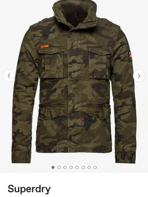 Camouflage vindjacka från Superdry - Snygg vindjacka från Superdry i klassiskt camouflage-mönster. Jackan har fyra stora fickor framtill, hög krage och coola detaljer som axelklaffar och patch på ärmen. Perfekt för dig som gillar streetstyle och vill sticka ut lite extra.