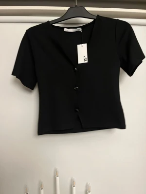 Svart kortärmad topp med knappar - En svart, croppad topp från 157 med korta ärmar och knappar framtill. Toppen har en enkel och stilren design med lätt v-ringad halsringning. Perfekt att matcha med jeans eller kjol för en clean look.