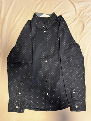  superclean prickig skjorta - Snygg svart skjorta med vita små prickar (polka dots) och klassisk krage. Skjortan har långa ärmar och vita knappar framtill samt vid ärmsluten. Perfekt för dig som vill ha en stilren men ändå lekfull look.