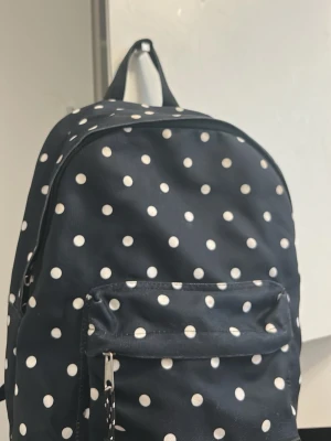 Svart prickig ryggsäck - Snygg svart ryggsäck med vita prickar och en praktisk framficka med dragkedja. Perfekt för skolan eller när du vill ha med dig det viktigaste. Väskan har ett klassiskt handtag upptill och justerbara axelband. Materialet känns slitstarkt och lätt att hålla rent.