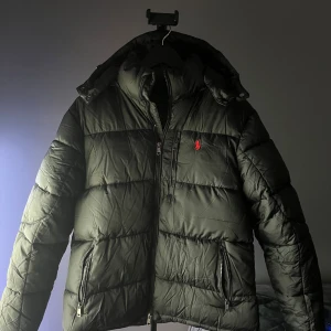 Svart pufferjacka från Polo Ralph Lauren - Säljer en svart pufferjacka från Polo Ralph Lauren med röd broderad logga på bröstet. Jackan har huva, två dragkedjefickor och quiltad design. Perfekt för kalla dagar och riktigt snygg till streetwear-stilen. Skicka bud!