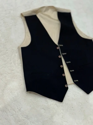 Svart klassisk kostymväst - Stilren svart väst med beige insida och fem knappar framtill. Västen har två diskreta fickor och en djup v-ringning. Perfekt för att lyfta en skjorta eller t-shirt till nästa nivå.