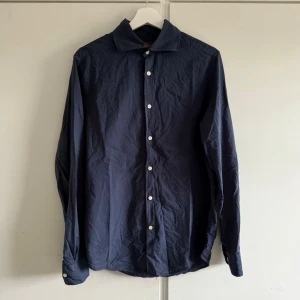 Marinblå skjorta Morris Slim Fit M - Mörkblå skjorta från Morris i slim fit-modell. Klassisk design med vita knappar och lång ärm. Tillverkad i bomull, perfekt för dig som gillar stilrena och enkla plagg. Snygg krage och diskret logga på bröstet. Lite urtvättad. Fri frakt