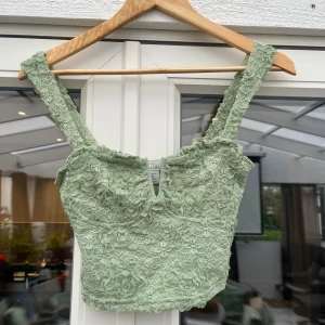 Grön spetsig korsettopp - Snygg ljusgrön korsettopp med blommig spets och croppad passform. Toppen har breda axelband och en liten v-ringning framtill. Perfekt för dig som vill sticka ut med en romantisk och trendig look.
