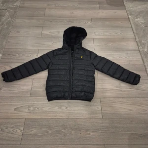 Svart pufferjacka från Lyle & Scott - Svart pufferjacka från Lyle & Scott med huva och gul logga på bröstet. Jackan är quiltad och har dragkedja framtill. Tillverkad i 100% polyester, perfekt för kyliga dagar. Enkel och stilren design som funkar till det mesta. Endast andvänd en gång och säljs pga att den ej passar storleken är då 12-13 år men sitter som S