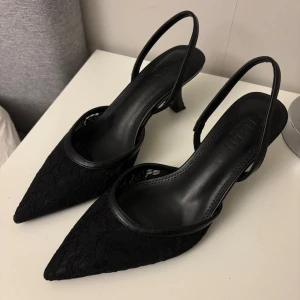 Svarta spetsiga pumps med låg klack - Svarta pumps från Simmi London med spetsig tå och låg klack. Skorna har en elegant spetsdetalj över hela ovandelen och tunna remmar runt hälen. Tillverkade i syntetmaterial med en klassisk och stilren look, perfekta för att lyfta vilken outfit som helst. Använd vid ett tillfälle!