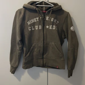 Olivgrön/millitärgrön hoodie från EDC by Esprit storlek M-XS - Olivgrön/millitärgrön hoodie från EDC by Esprit med broderad text på bröstet och patch på ärmen. Tröjan har dragkedja, huva med snörning och mysigt teddyfoder på insidan. Perfekt för kyliga dagar och har två stora fickor framtill. Storlek M men funkar även till barn så M-XS.
