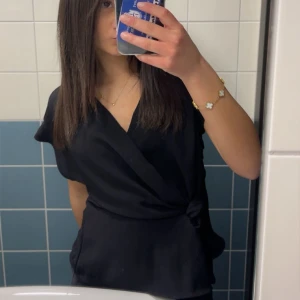 Svart omlottopp med knyt - Snygg svart omlottopp med knyt i sidan och v-ringad hals. Toppens material är mjukt och har en lätt glans, vilket ger en stilren look. Perfekt att matcha med jeans eller kjol för en trendig outfit. Blusen är ifrån Zara och i mycket bra skick 