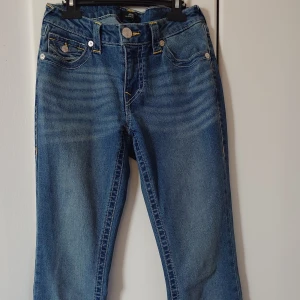 True Religion blå jeans med kontrastsömmar - Säljer ett par blå jeans från True Religion med tydliga kontrastsömmar och snygga bakfickor med lock och dekorativa knappar. Klassisk femficksmodell med gylf och knapp, tillverkade i bomullsmix för skön stretch. Perfekta för dig som gillar streetwear vibes.