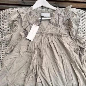 Grön volangblus från Neo Noir - Supersöt blus från Neo Noir i färgen smoke green. Blusen har volangdetaljer längs axlar och bröst, v-ringning och fina broderade partier. Luftig och lätt i bomull, perfekt för vår och sommar. Modellen heter Jayla S Voile Top . Det är samma färg som första bilden men det blev väldigt dåligt ljus, skriv för fler bilder. Blusen är helt ny och aldrig andvänd, lappen är kvar.