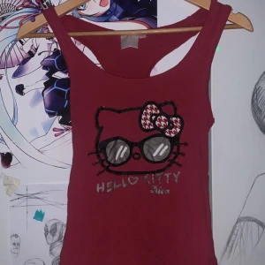 Röd Hello Kitty linnetopp - Snygg röd linnetopp med Hello Kitty-tryck framtill, där katten har stora solglasögon och en rosett med rutigt mönster. Toppen är ärmlös och har brottarrygg, perfekt för en lekfull och avslappnad stil. Det finns några små hål. Alla hålen fick inte plats på bild. Vrt tyvärr inte storlek eller märke men skulle säga att den passar M. Axelbanden är långa.