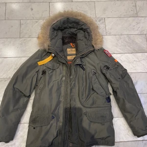 Parajumpers grön parkajacka med päls - Säljer en grön Parajumpers parkajacka med pälsfodrad huva och flera fickor. Jackan har ikoniska PJS-patchar på ärmen, gul detalj vid axeln och dragkedja framtill. Slim fit-modell från Masterpiece Series, perfekt för kalla dagar.