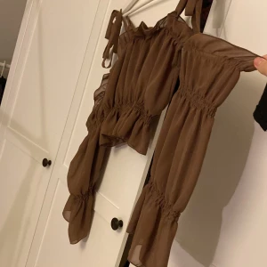 Brun blus med volang från Buch Copenhagen - Supersöt brun blus från Buch Copenhagen med volangdetaljer och smock i midjan och vid ärmslut. Blusen har långa ärmar, knytband på axlarna och är gjord i ett tunt, flowy material som ger en luftig känsla. Perfekt för dig som gillar romantiska vibes.