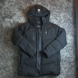 Svart dunjacka Sail Racing Antarctica - Svart dunjacka från Sail Racing Antarctica med huva, två bröstfickor med dragkedja och snygga detaljer på ärmen. Jackan är vadderad och har en stilren look, perfekt för kalla dagar. Klassisk passform och diskret logga på bröstet och ärmen.