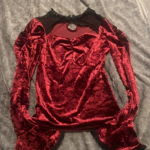 Vinröd sammets tröja med spets - Supersnygg vinröd sammets tröja med långa ärmar och svart spetsdetalj vid halsen. Klänningen har en liten rosett framtill, nyckelhålsöppning i nacken och volang vid ärmsluten. Perfekt för dig som vill sticka ut med en lyxig och unik look.