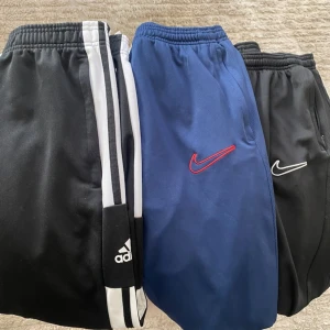 Adidas och Nike mjukisbyxor - Tre par mjukisbyxor, ett svart med vita ränder från Adidas och två från Nike, ett blått med röd logga och ett svart med vit logga. Alla har resår i midjan och är perfekta för en sportig och avslappnad stil.
