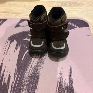 Bruna vinterkängor med kardborre stlr 20 (barn) - Säljer ett par bruna vinterkängor från Reima med grov svart sula och kardborreband. Utsidan är i slitstark syntet och mocka, fodret är svart och mjukt. Dekor med gula stjärnor och krona på sidan, samt reflexdetaljer. Perfekt för kalla dagar!