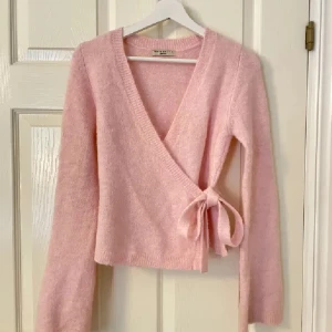 Rosa omlottkofta från Gina Tricot - Superfin ljusrosa omlottkofta från Gina Tricot med knyt i sidan. Koftan är stickad och har långa ärmar samt v-ringning. Perfekt att styla med ett linne under för en söt och trendig look.