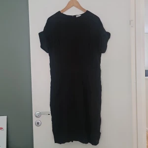 Svart klänning från Filippa K XL - En svart, stilren klänning från Filippa K i storlek XL. Klänningen har korta ärmar, rund halsringning och dragkedja bak. Den är knälång och har en rak, enkel passform som ger en clean look. Perfekt för dig som gillar minimalistisk och tidlös stil.