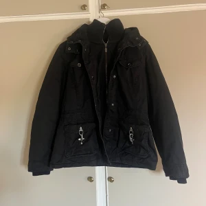 Svart parkajacka med dragkedja - Svart parkajacka med hög ribbad krage, dragkedja framtill och stora fickor med metallspännen. Jackan har långa ärmar med ribbade muddar och flera snygga detaljer som tryckknappar och justerbar midja. Perfekt för kyligare dagar.