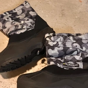 Blåsvarta vinterstövlar från Bluewear - Tuffa vinterstövlar från Bluewear med svart och grått camouflagemönster upptill och grov sula för extra grepp. Ovandelen är i slitstark syntet och nederdelen i kraftig gummi. Dragsko upptill för att hålla snön ute. Perfekta för kalla dagar.
