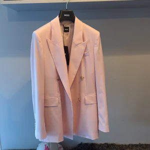 Ljusrosa kavaj från BOSS - Snygg  ljusrosa kavaj från BOSS med dubbelknäppning och spetsiga slag. Kavajen har två framfickor med lock och coola marmorerade knappar. Materialet är slätt och har en lätt glansig finish som ger en exklusiv vibe. Perfekt för dig som vill sticka ut med stil. 