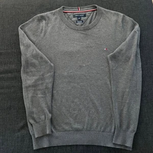 Grå tröja från Tommy Hilfiger - Snygg grå tröja från Tommy Hilfiger med rund halsringning och diskret logga på bröstet. Tröjan har långa ärmar och ribbade muddar vid ärmslut och nederkant. Perfekt för lager-på-lager under kyliga dagar.