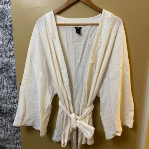 Vit omlott overshirt från LinDEX - Luftig och stilren vit overshirt från LinDEX med breda ärmar och knytband i midjan. Jackan har en krinklad struktur och är tillverkad i ett lätt bomullsmaterial. Perfekt för lager-på-lager och ger en chill vibe till din outfit.