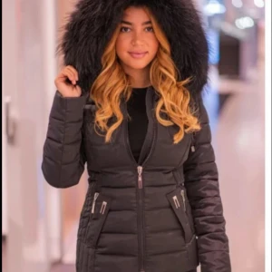 Svart pufferjacka med fuskpäls - Snygg svart pufferjacka med stor huva och fluffig fuskpäls. Jackan har dragkedja framtill, två snedställda fickor med dragkedjor och markerad midja för en schysst siluett. Perfekt för kalla dagar när du vill hålla stilen. Från garoff!!