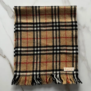 Rutig halsduk från Burberrys i ull - Klassisk rutig halsduk från Burberrys i 100% Shetlandsull. Beige bas med svarta, vita och röda rutor samt fransar i ändarna. Perfekt accessoar för att lyfta din outfit med en tidlös brittisk stil.