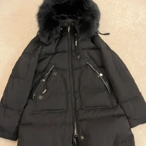 Svart dunjacka från Cedrico Norway XS - Snygg svart dunjacka från Cedrico Norway i storlek XS. Jackan har fluffig fuskpäls runt huvan, flera dragkedjor och stora fickor med detaljer i skinn. Perfekt för kalla dagar med sin tjocka vaddering och stilrena look. Har köpt jackan för 5500 