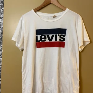 Vit Levi's t-shirt med logga - Vit t-shirt från Levi's med klassisk stor logga i blått, svart och rött framtill. T-shirten har rund halsringning och korta ärmar. Tillverkad i mjuk bomull och har en avslappnad passform som funkar till allt.