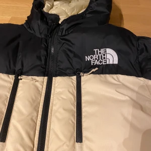 Beige och svart pufferjacka The North Face - Snygg pufferjacka från The North Face i beige och svart med huva och flera dragkedjeförsedda fickor. Jackan har broderad logga på bröstet och är vadderad för extra värme. Perfekt för kalla dagar och har en modern, sportig look.