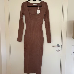 Brun stickad klänning från NAKD - Säljer denna snygga o mjuka klänning från NAKD då den inte var rätt storlek på mig! Den är helt oanvänd, endast testad. Klänningen har en slits på höger sida och detaler på framsidan. Storlek M, går ner till mitten av vaden ungefär på mig (170 cm). ps. Klänningen är lite ljusare i färgen i verkligheten än på bilderna