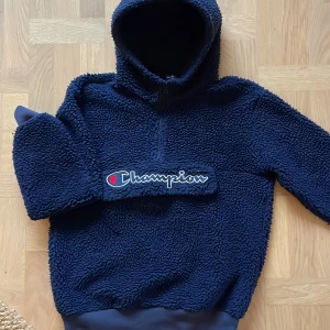 Mörkblå fleece hoodie från Champion - Mysig mörkblå hoodie från Champion i teddyfleece med stor broderad logga framtill. Tröjan har huva, half zip och långa ärmar med muddar. Perfekt för kalla dagar när du vill vara både varm och snygg. Praktisk ficka framtill. 