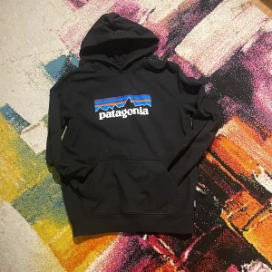 Svart Patagonia hoodie med logga - Svart hoodie från Patagonia med stor färgglad logga på bröstet. Klassisk känguruficka framtill och huva. Tröjan är i bomull och polyester, perfekt för chill dagar eller när du vill ha en avslappnad stil.