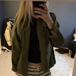 Olivgrön overshirt från Gina Tricot - Säljer en olivgrön jeansjacka från Gina Tricot med klassisk krage och flera stora fickor framtill. Jackan har lång ärm, knappar och är tillverkad i mjuk bomull. 