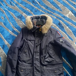 Mörkblå Parajumpers parkajacka - Säljer en mörkblå parkajacka från Parajumpers med päls på huvan och flera praktiska fickor. Jackan har quiltat foder, dragkedja och knappar framtill samt snygga detaljer som patch på ärmen och gul rem vid kragen. Perfekt för kalla dagar.
