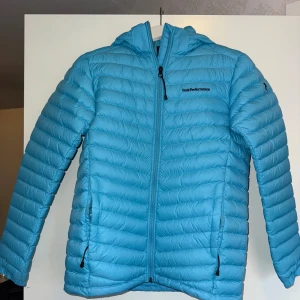 Blå dunjacka Peak Performance  - Snygg blå dunjacka från Peak Performance med huva och dragkedja framtill. Jackan har quiltad design, två fickor med dragkedja och broderad logga på bröstet. Perfekt för kyliga dagar. Storlek 170