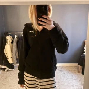 Svart Nike hoodie med dragkedja - Svart hoodie från Nike med vit logga på bröstet. Tröjan har dragkedja framtill, huva med snörning och två fickor. Perfekt för chill dagar eller när du vill ha en sportig look. Mjuk och skön, passar till allt. Använd ett fåtal gånger! 