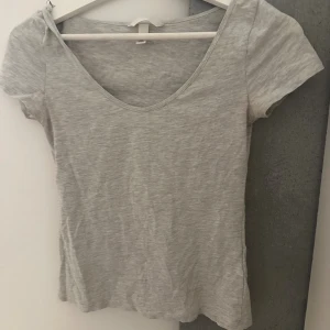 Ljusgrå t-shirt från H&M i stl S - En basic ljusgrå t-shirt från H&M med rundad, djup urringning och korta ärmar. Modellen är figurnära och tillverkad i mjuk bomullsmix.