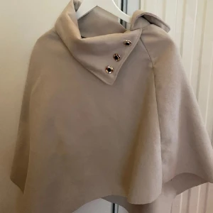 Beige poncho med knappar - Snygg beige poncho med hög krage och tre dekorativa knappar vid axeln. Ponchon har en lös och svepande passform som ger en chill och snygg look. Passar för lager-på-lager-stil under kyliga dagar🌸