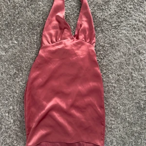 Rosa halterneck klänning PrettyLittleThing - Supersnygg kort halterneck klänning i satinliknande material från PrettyLittleThing. Klänningen är rosa, har djup v-ringning och knyt i ryggen för en extra cool look. Perfekt för dig som vill sticka ut med en trendig och feminin stil.