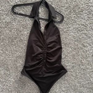 Svart halterneck body med knappar - Snygg svart body med halterneck och knyt i nacken. Framsidan har dekorativa knappar och lätt rynkade detaljer som ger en cool look. 