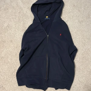 Marinblå hoodie från Polo Ralph Lauren - Säljer en marinblå hoodie från Polo Ralph Lauren med dragkedja och huva. Tröjan har klassisk röd logga broderad på bröstet, kängurufickor och långa ärmar. Perfekt för chill dagar eller när du vill ha en clean och stilren look.